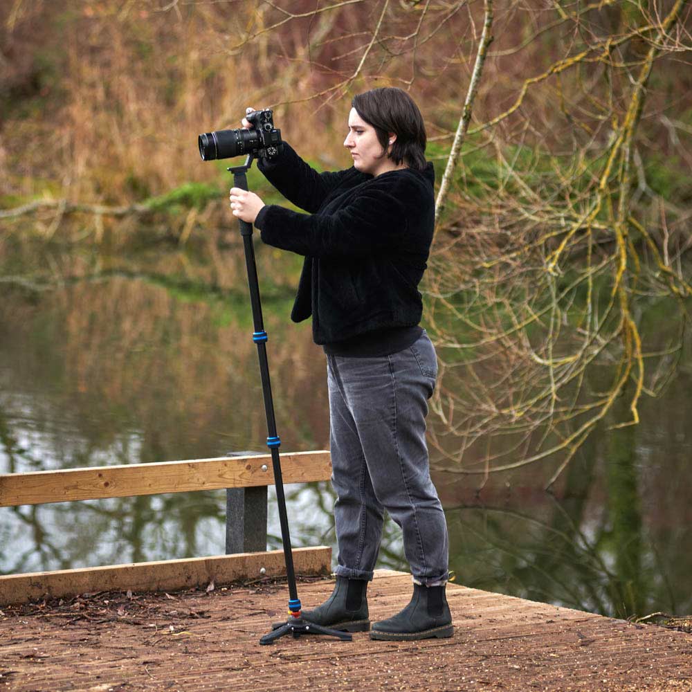 Person holding a camera on a monopod with tripod feet by a body of water