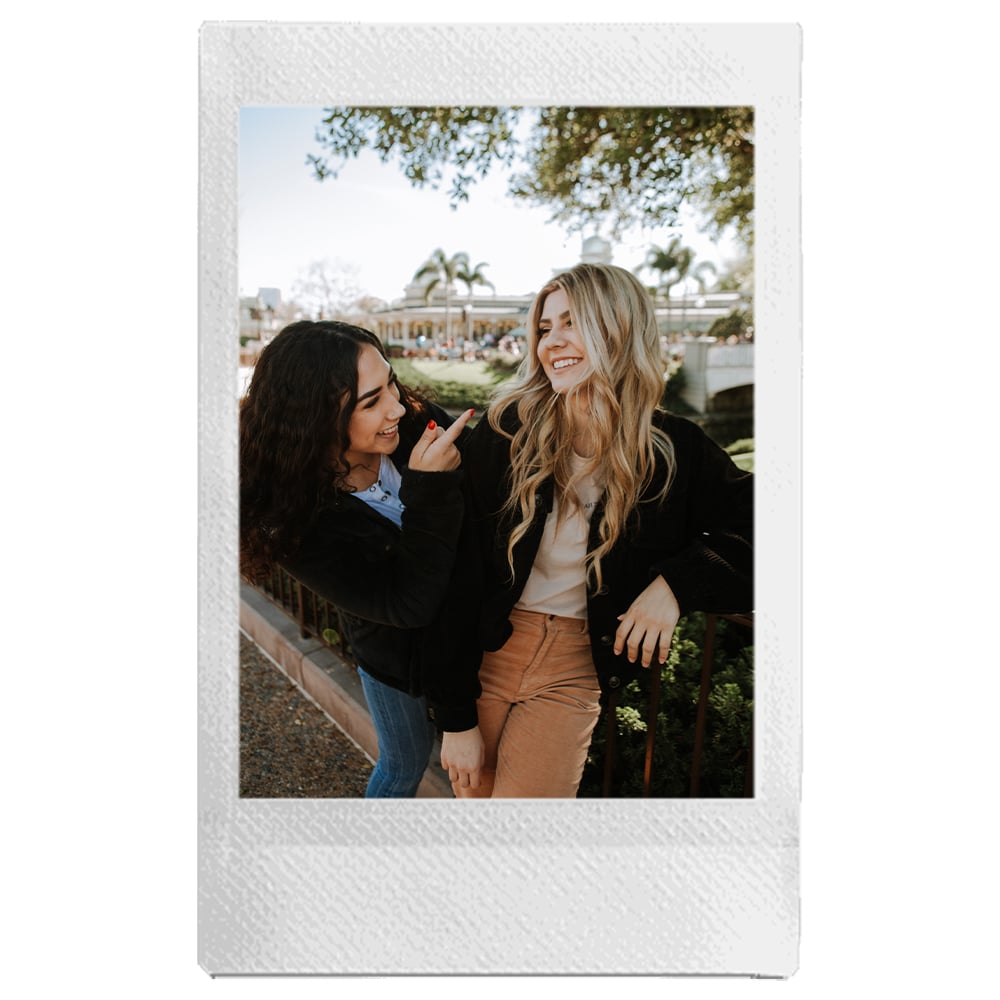 Two women standing outdoors, smiling and holding hands, with a blurred background.