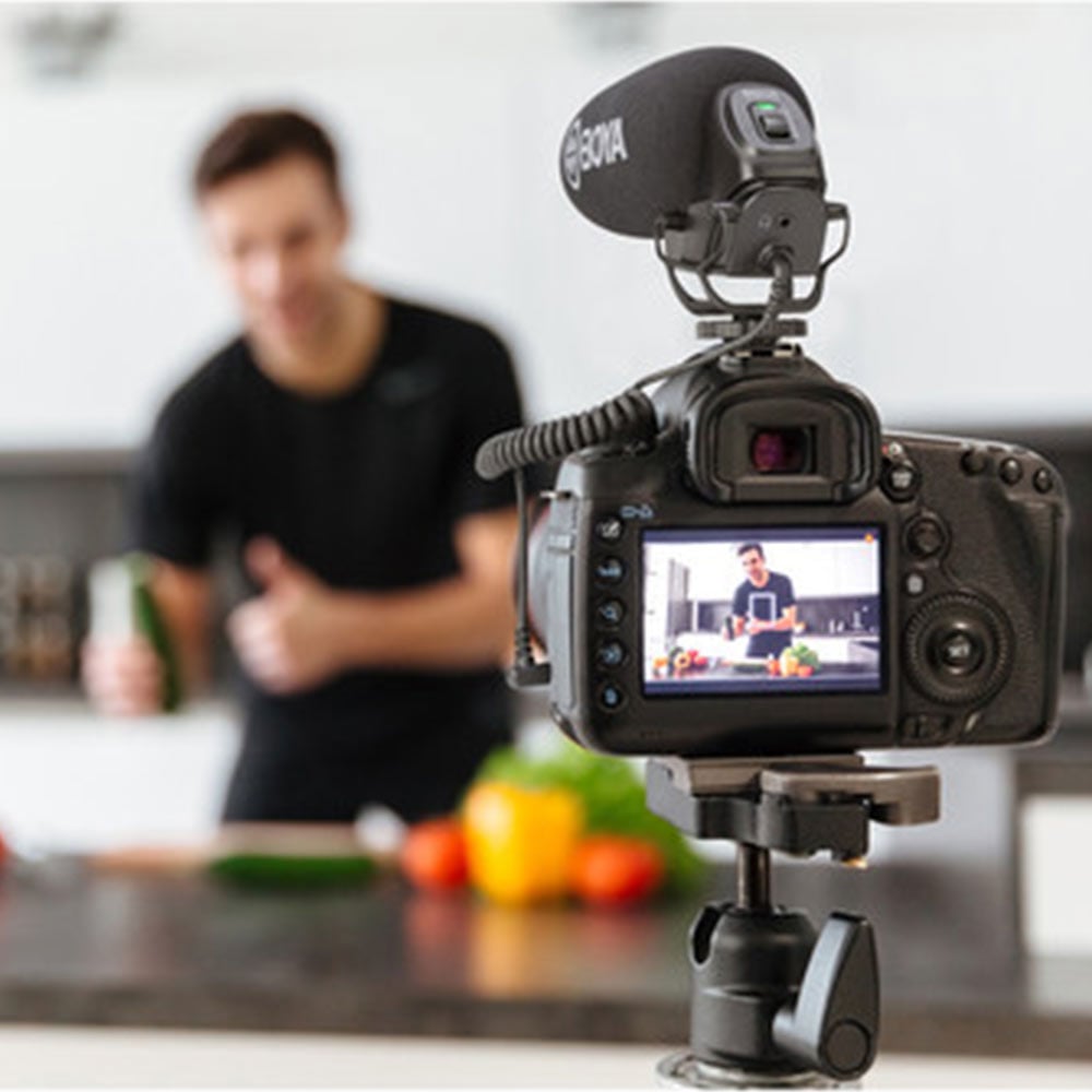 Camera on a tripod recording a person in a kitchen setting with vegetables.