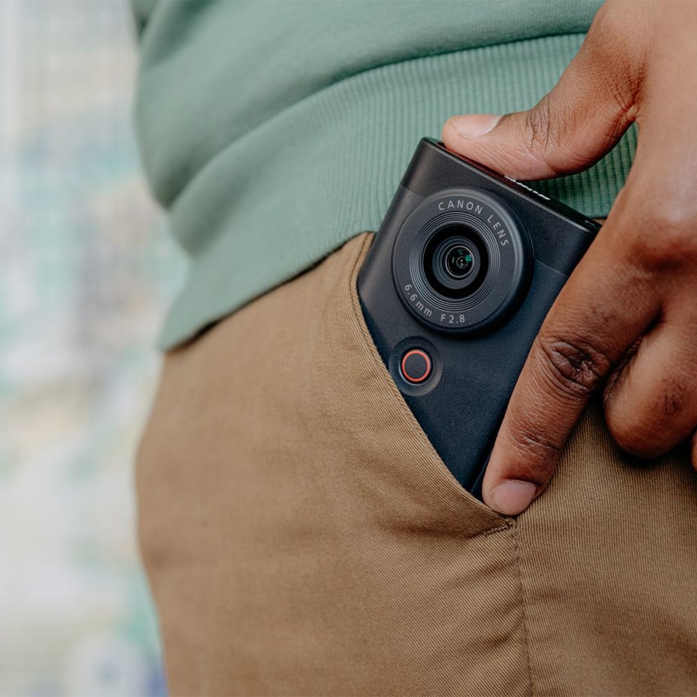 Canon PowerShot V10 vlog camera kit in silver — compact rectangular camera with wide-angle lens, flip-up screen, and integrated mini tripod stand.