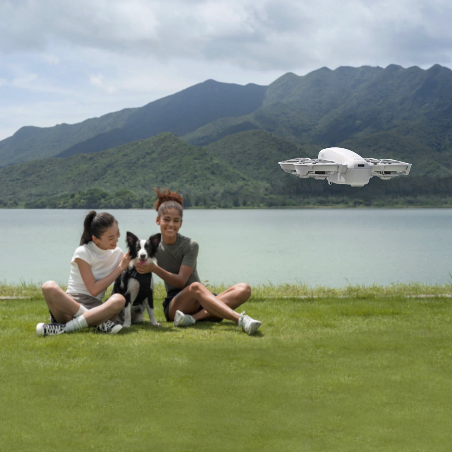 Two people sitting on grass with a dog, a flying drone in the sky, and mountains in the background.