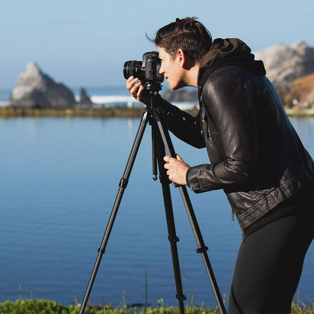 Person using a camera on a tripod by a body of water with mountains in the background