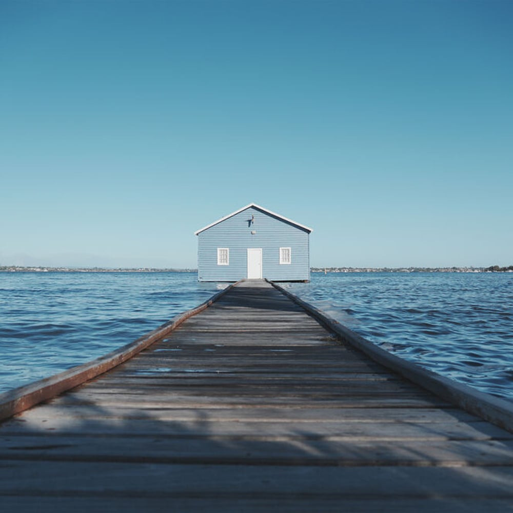 Blue boathouse at the end of a wooden pier over calm water with clear blue sky.