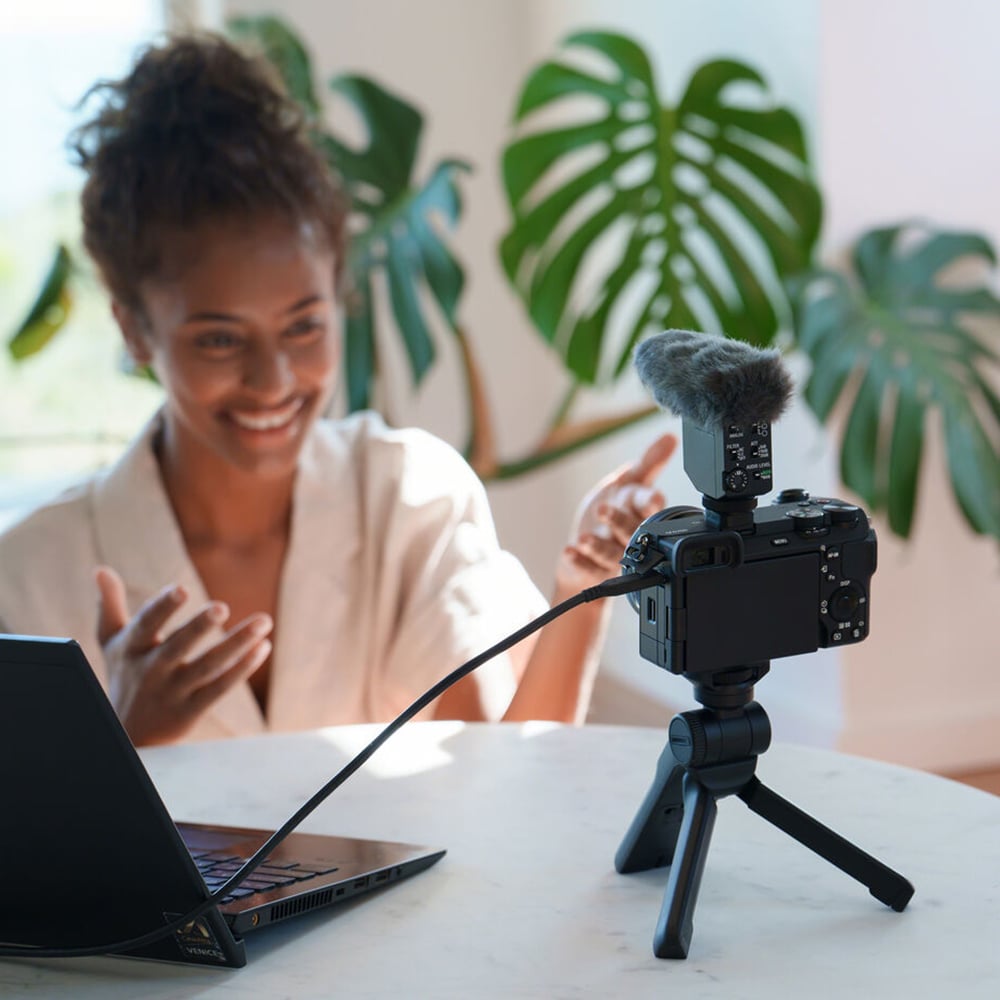 Woman recording a video with a camera on a tripod in front of her, with plants in the background.